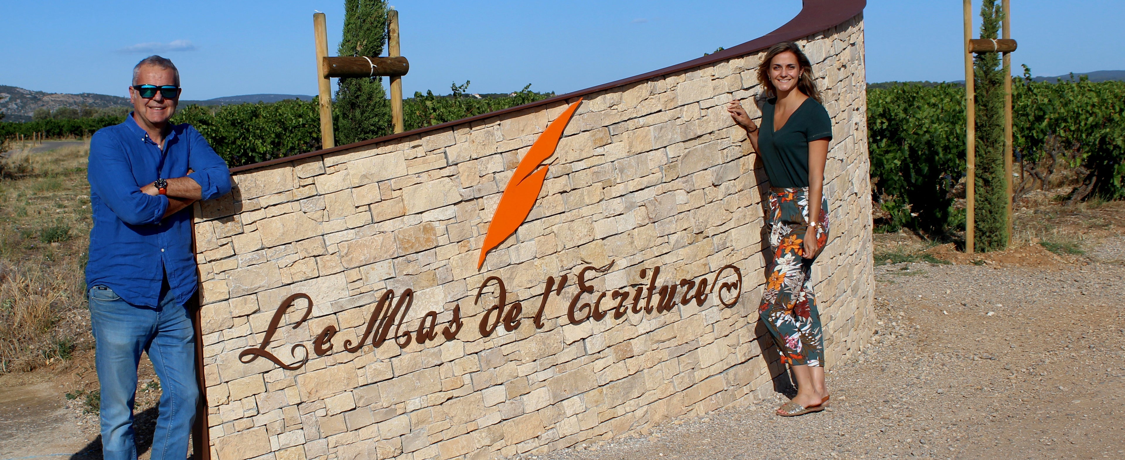 Pascal et Léa Fulla posent fièrement devant l'écriteau 'Mas de l'Écriture' à l'entrée de leur domaine viticole. Cette photo capture l'esprit familial et l'engagement envers la viticulture durable dans les Terrasses du Larzac, Languedoc.